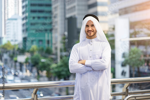 A Caucasian Arab man standing on a bridge platform with arms across the chest posture, wearing rational arab garment disdasha and thobe, within the city center, nature and busy road in the background photo