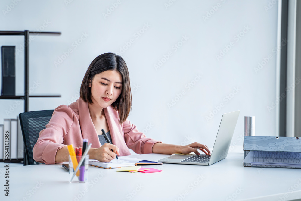 Obraz premium beautiful female asian worker using laptop computer typing on keyboard and writing on notebook, work in an office with folder files sticky notes for ideas information, wearing pink formal clothing.