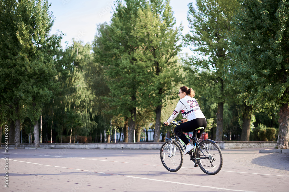 Sep 7, 2019-Ternopil/Ukraine:Young brunette woman, wearing black leggings and white pullover, riding a bike in city town park with green trees in summer.Portrait of girl, cycling. Healthy life concept