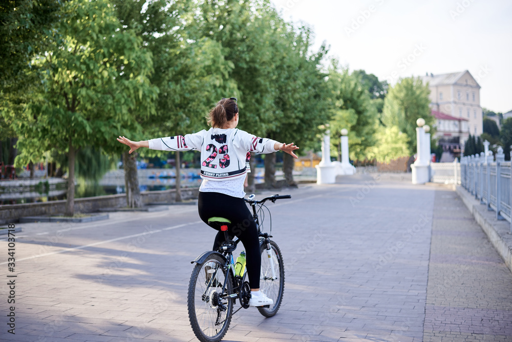Sep 7, 2019-Ternopil/Ukraine:Young brunette woman, wearing black leggings and white pullover, riding bike in town park in summer. Portrait of girl, cycling, doing acrobatic tricks,lifting her arms up.