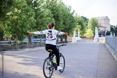 Sep 7, 2019-Ternopil/Ukraine:Young brunette woman, wearing black leggings and white pullover, riding bike in town park in summer. Portrait of girl, cycling, doing acrobatic tricks,lifting her arms up. © Natalia