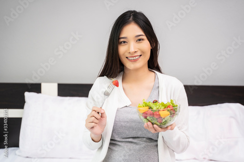 Beautiful asian pregnant woman holding a bowl of salad of vegetables and fruits, using a fork eating a strawberry. Eating healthy having lunch sitting on the white bed, relaxing and resting in bedroom