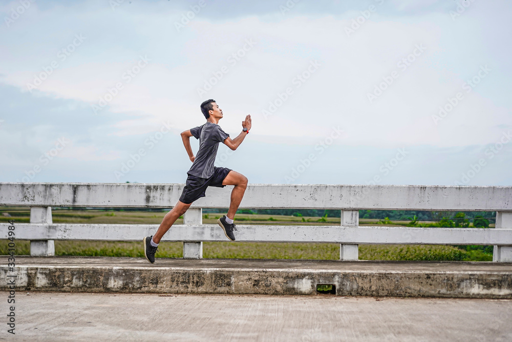 asian teenager running along the bridge in sprinting action, exercising or practicing for a marathon race, wearing shirt, short and running shoes, with tree, nature and cloudy sky in the background