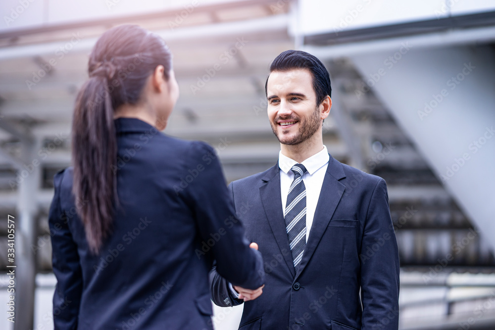 caucasian businessman smiling and standing casually shaking hands with a beautiful asian businesswoman in greetings, outdoor within the city district with urban architectural structure in background