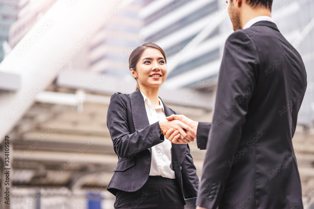 asian businesswoman smiling shaking hands greeting and social interaction with a tall handsome caucasian businessman, outdoor within the city district with urban architectural structure in background asian businesswoman smiling shaking hands greeting and social interaction with a tall handsome caucasian businessman, outdoor within the city district with urban architectural structure in background