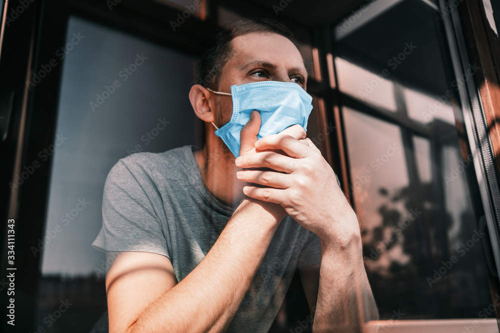 man in quarantine because of a virus sits at home in a mask and looks out the window on a sunny day..Coronavirus concept. COVID-19