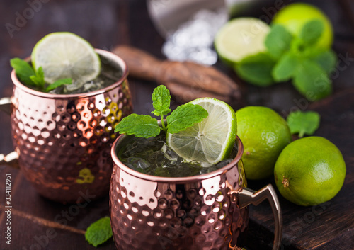 Moscow mule cocktail in a copper mug with lime and mint and wooden squeezer on dark wooden background.