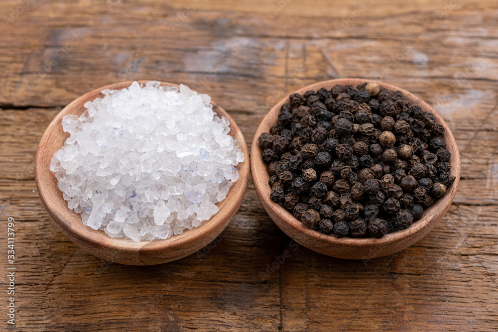 Coarse crystal salt and whole black peppercorns in small wooden bowls on a rustic wooden background Coarse crystal salt and whole black peppercorns in small wooden bowls on a rustic wooden background