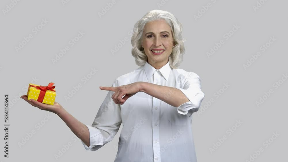 Happy old lady holding gift box on her palm. Positive smiling lady wearing white shirt giving present. Grey isolated background. Happy old lady holding gift box on her palm. Positive smiling lady wearing white shirt giving present. Grey isolated background.