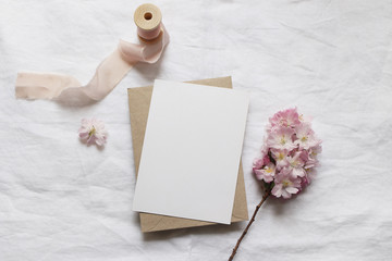 Wedding stationery mock-up scene. Blank vertical greeting card, envelope on linen tablecloth background with pink blossoming cherry tree branches and ribbon. Feminine still life composition. Flat lay.