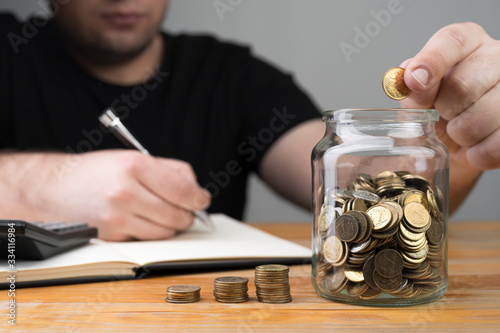 Coins in a jar, stacks of change and young caucasian man dropping money and writing in a notebook. Tax forms, counting savings, money collecting or accounting abstract concept.