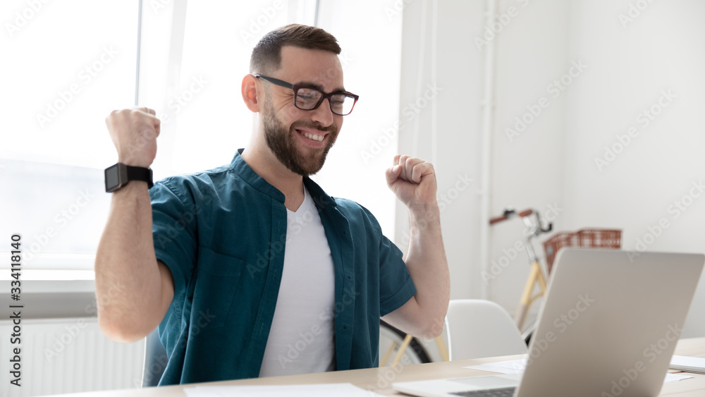 Overjoyed young male employee in glasses sit at office desk feel euphoric read good promotion news on laptop, excited man worker triumph win online lottery on computer, reward, luck concept
