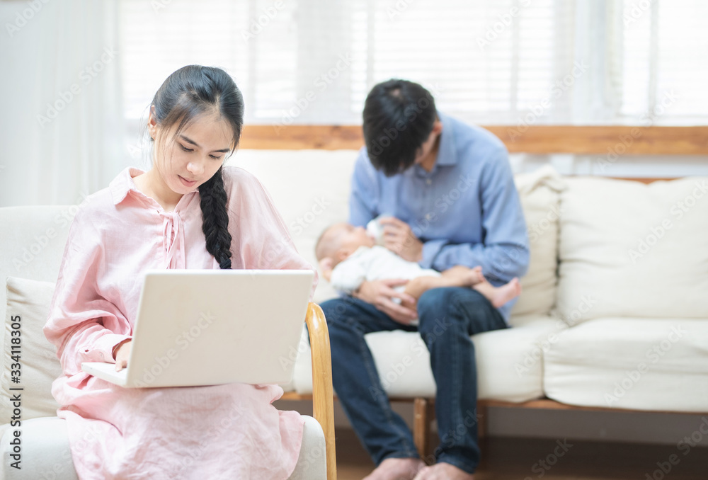 Young asian mother using laptop computer in living room at home with blurred of father acting mom feeding milk bottle in background his son baby 1 year old, work at home and social distancing concept. Young asian mother using laptop computer in living room at home with blurred of father acting mom feeding milk bottle in background his son baby 1 year old, work at home and social distancing concept.