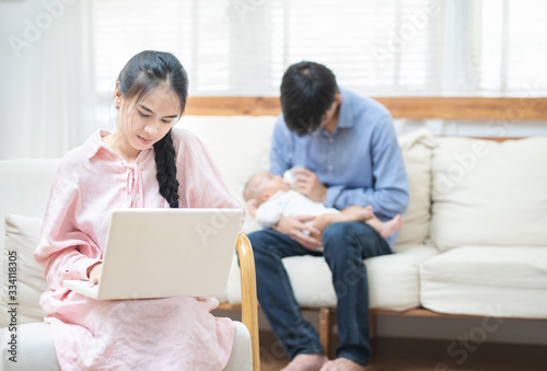 Young asian mother using laptop computer in living room at home with blurred of father acting mom feeding milk bottle in background his son baby 1 year old, work at home and social distancing concept.