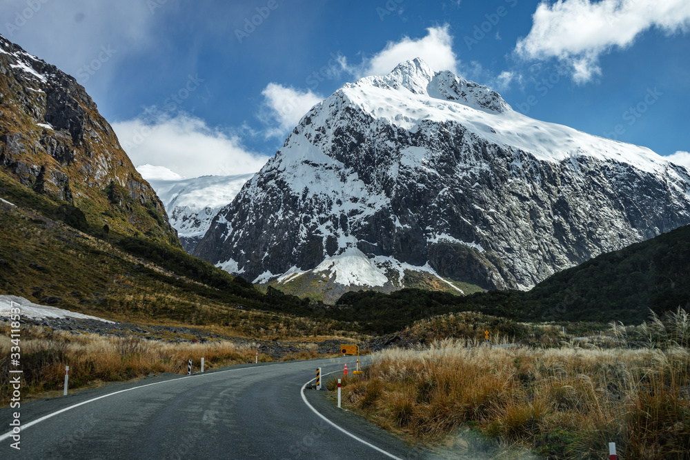 Splendid image of the SH94 road towards Milford Sound surrounded by greenery with snow capped mountains in the background taken on a sunny winter day, New Zealand