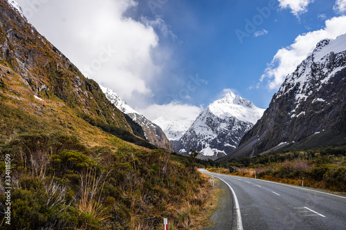 Splendid image of the SH94 road towards Milford Sound surrounded by greenery with snow capped mountains in the background taken on a sunny winter day, New Zealand