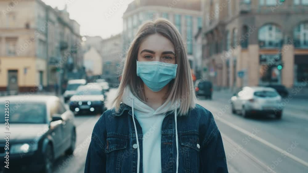 Young caucasian woman in medical mask standing in city street looking to camera road with cars on background Concept of health and safety life COVID-19 coronavirus virus protection pandemic in world