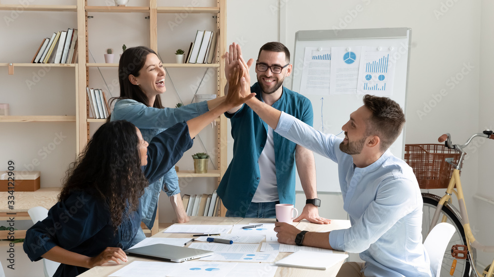 Overjoyed diverse businesspeople gather in office gave high five celebrate shared victory or win, happy multiracial colleagues coworkers join hands engaged in teambuilding activity at meeting - obrazy, fototapety, plakaty 