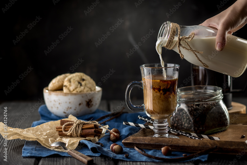 female hand pouring milk into glass of black coffee with cookies and cinnamon sticks on black background
