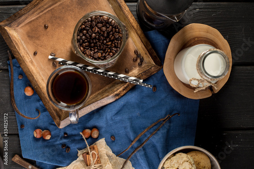 Cup of coffee with coffee beans, spoon, cinnamon sticks, bottle of milk and cookies on blue background
