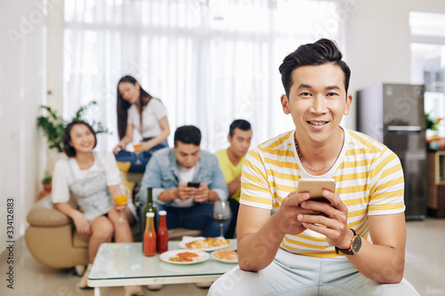 Smiling Vietnamese young man texting and looking at camera when his friends eating and drinking in background at house party