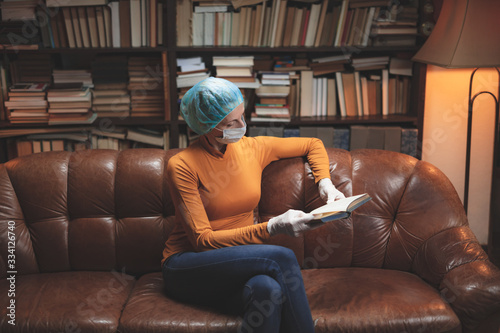 Woman with protective antiviral mask sitting at home in isolation / quarantine and reading book.