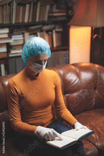 Woman with protective antiviral mask sitting at home in isolation / quarantine and reading book.