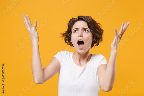 Shocked irritated young brunette woman girl in white t-shirt posing isolated on yellow orange background in studio. People lifestyle concept. Mock up copy space. Spreading hands screaming, looking up.