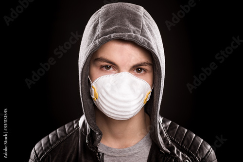 Concept of coronavirus quarantine. Teen boy wearing medical face mask to health protection from influenza virus, on black background. COVID-19 - home isolation. Emotional portrait of teenager in hood