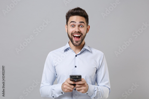 Excited young unshaven business man in light shirt posing isolated on grey background in studio. Achievement career wealth business concept. Mock up copy space. Using mobile phone, typing sms message.