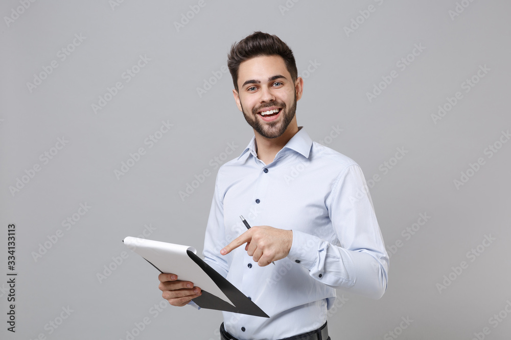 Cheerful young unshaven business man in light shirt isolated on grey background. Achievement career wealth business concept. Mock up copy space. Point index finger on clipboard with papers document. Cheerful young unshaven business man in light shirt isolated on grey background. Achievement career wealth business concept. Mock up copy space. Point index finger on clipboard with papers document.