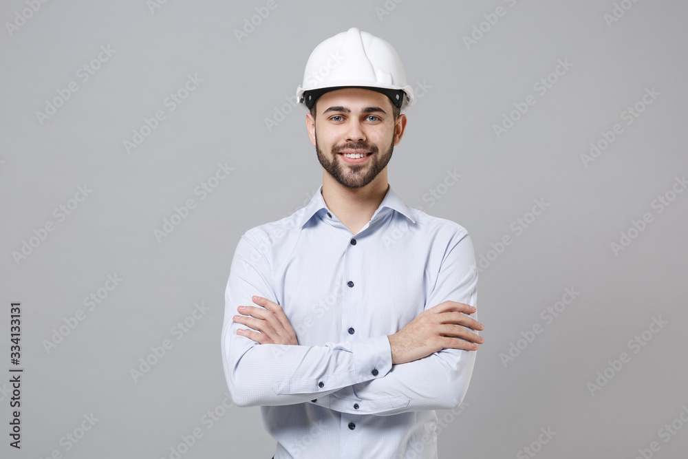 Smiling young unshaven business man in light shirt protective construction helmet isolated on grey background. Achievement career wealth business concept. Mock up copy space. Holding hands crossed. Smiling young unshaven business man in light shirt protective construction helmet isolated on grey background. Achievement career wealth business concept. Mock up copy space. Holding hands crossed.