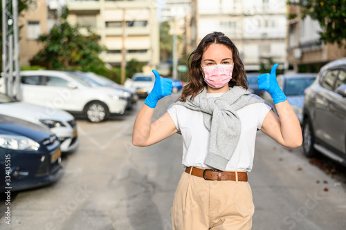 Beautiful girl in protective gloves and a medical mask shows like, thumbs up. She is standing on the street. The concept of self-defense and prevention of viruses, epidemics. Urban view. Close-up.