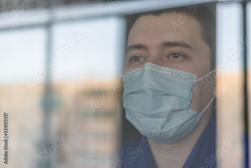 A young man in a medical mask remains isolated at home for self-quarantine. The concept of home quarantine, prevention of COVID-19, the situation with the coronavirus outbreak. A person who works from