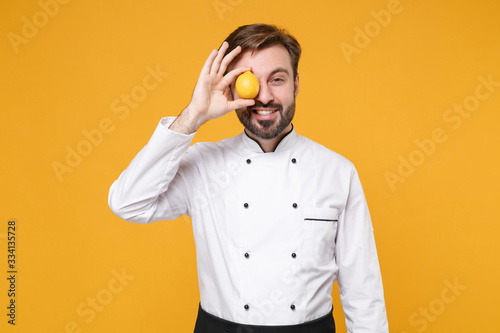 Smiling young bearded male chef cook or baker man in white uniform shirt posing isolated on yellow wall background studio portrait. Cooking food concept. Mock up copy space. Covering eye with lemon.