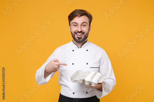 Cheerful young bearded male chef cook or baker man in white uniform shirt posing isolated on yellow background in studio. Cooking food concept. Mock up copy space. Pointing index finger on egg tray.