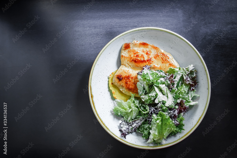 The main dish. Grilled chicken breast with a crust on a plate with mashed potatoes and green salad on a black background. Top view. The main dish. Grilled chicken breast with a crust on a plate with mashed potatoes and green salad on a black background. Top view.
