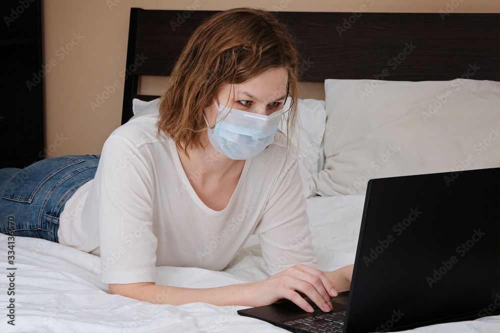 Woman wearing medical mask working lying on a bed with laptop computer. Corona virus quarantine, isolation period covid - 19. Social distancing
