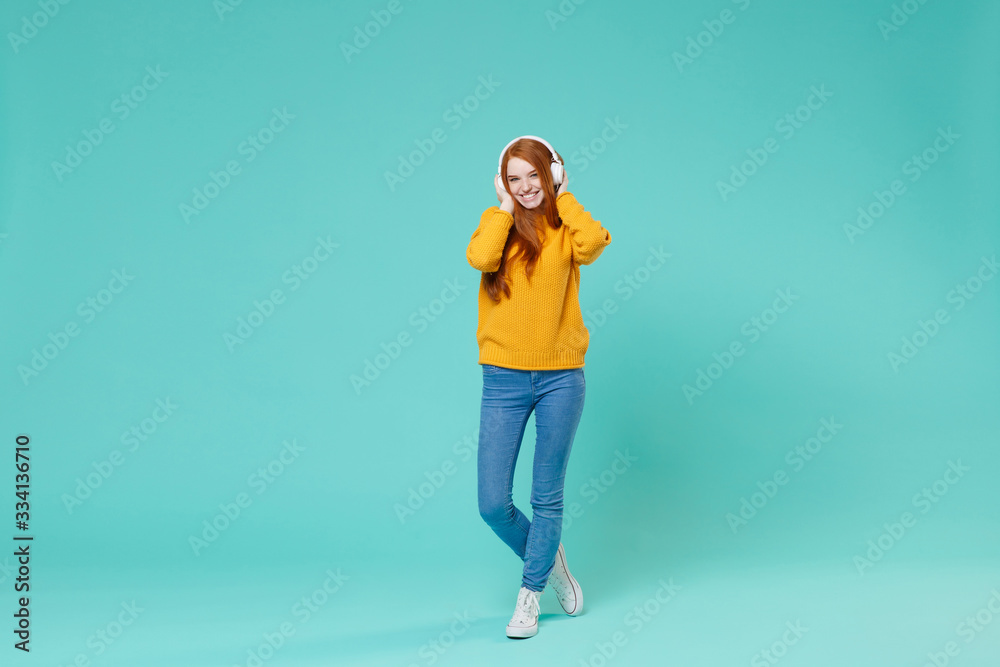 Pretty young redhead girl in yellow knitted sweater posing isolated on blue turquoise background studio portrait. People emotions lifestyle concept. Mock up copy space. Listen music with headphones. Pretty young redhead girl in yellow knitted sweater posing isolated on blue turquoise background studio portrait. People emotions lifestyle concept. Mock up copy space. Listen music with headphones.