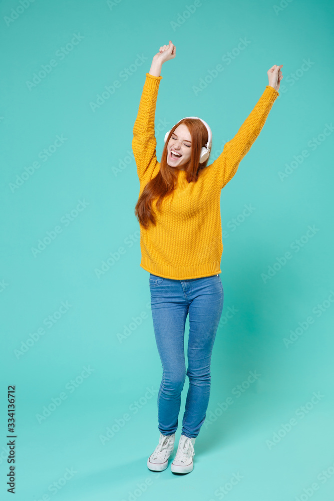 Funny young redhead girl in yellow knitted sweater posing isolated on blue turquoise background studio portrait. People lifestyle concept. Mock up copy space. Listen music with headphones, dancing. Funny young redhead girl in yellow knitted sweater posing isolated on blue turquoise background studio portrait. People lifestyle concept. Mock up copy space. Listen music with headphones, dancing.