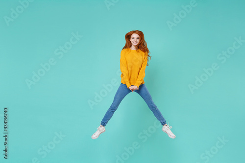 Smiling young redhead woman girl in yellow knitted sweater posing isolated on blue turquoise background studio portrait. People emotions lifestyle concept. Mock up copy space. Jumping spreading legs.