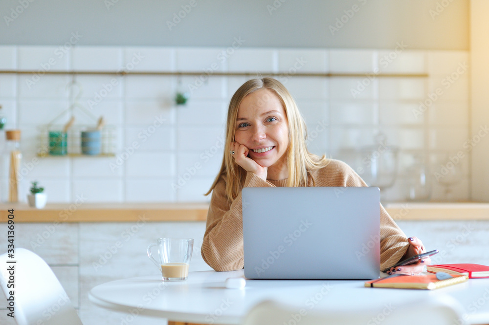 Lovely blonde woman working with her laptop while sitting in the kitchen in her apartment Lovely blonde woman working with her laptop while sitting in the kitchen in her apartment