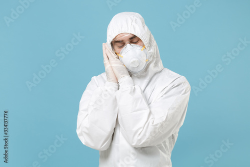 Man in white protective suit respirator mask sleep with folded hands under cheek isolated on blue background studio. Epidemic pandemic rapidly spreading coronavirus 2019-ncov medicine virus concept.