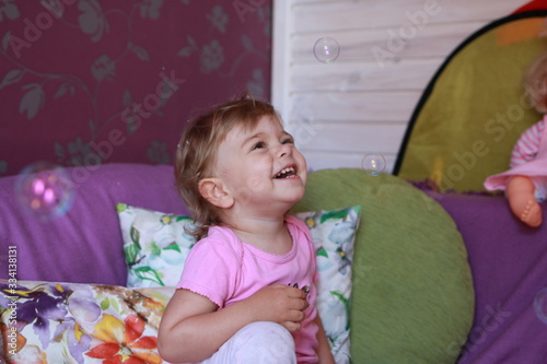 Cute little Caucasian girl one year old plays in her room with soap bubbles, laughs merrily, close-up. Childhood, leisure, games.