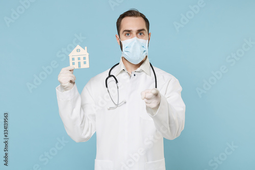 Male doctor man in medical gown face mask gloves isolated on blue background. Epidemic pandemic coronavirus 2019-ncov sars covid-19 flu virus. Hold house recommending stay home point finger on camera.