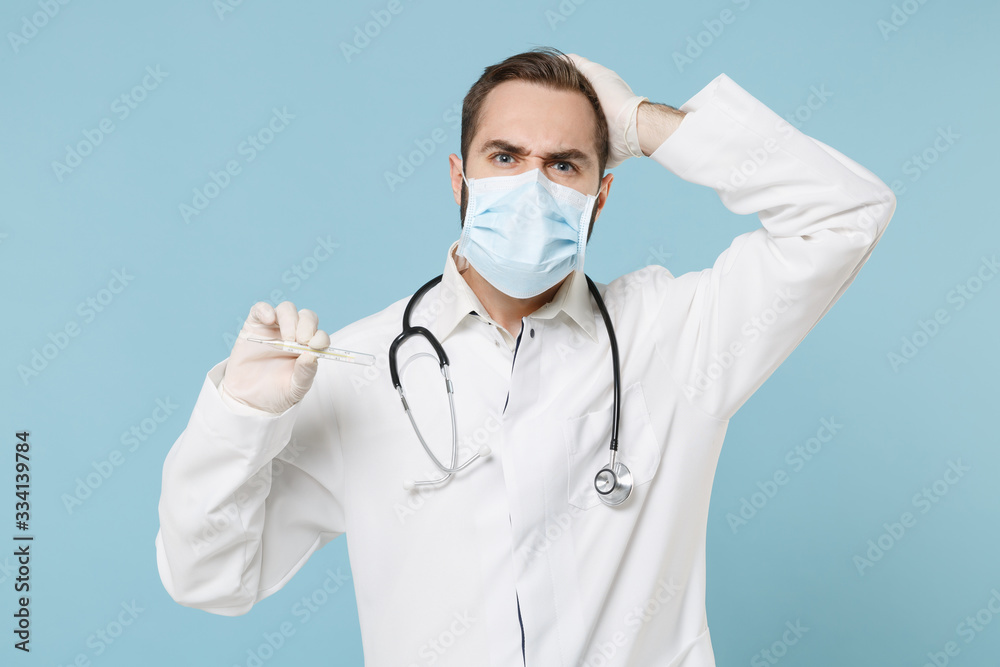 Preoccupied male doctor man in medical gown face mask gloves isolated on blue background. Epidemic pandemic coronavirus 2019-ncov sars covid-19 flu virus concept. Hold thermometer put hand on head. Preoccupied male doctor man in medical gown face mask gloves isolated on blue background. Epidemic pandemic coronavirus 2019-ncov sars covid-19 flu virus concept. Hold thermometer put hand on head.