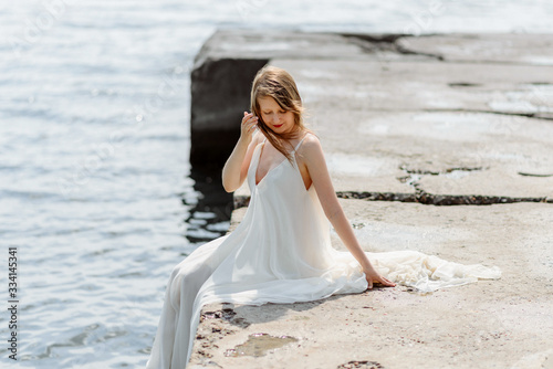 A young beautiful girl in a long milk-colored dress walks along the beach and pier against the background of the sea.