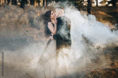 A loving bridegroom in a black cardigan and a cute bride in an expensive dress are holding a pyrotechnic device with smoke in their hands and hugging. Wedding portrait of the newlyweds.