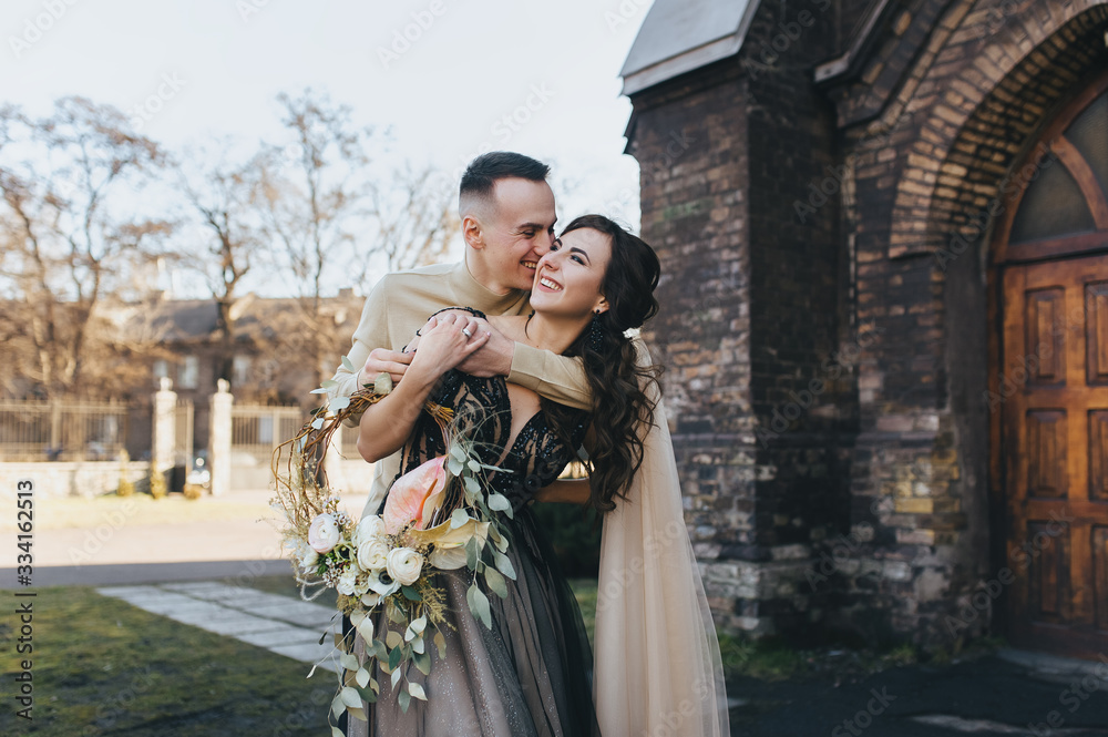 Stylish groom and sweet bride in an expensive dress hug and laugh, fooling around against the background of an old church. Wedding portrait of lovers newlyweds. Tender photography, concept.