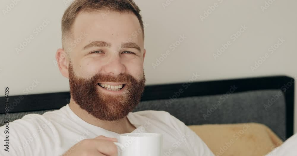 Close up view of handsome caucasian man in 30s smelling coffee . Portrait of bearded guy in 30s enjoing good morning while lying in bed and smiling to camera. Home background. Close up view of handsome caucasian man in 30s smelling coffee . Portrait of bearded guy in 30s enjoing good morning while lying in bed and smiling to camera. Home background.